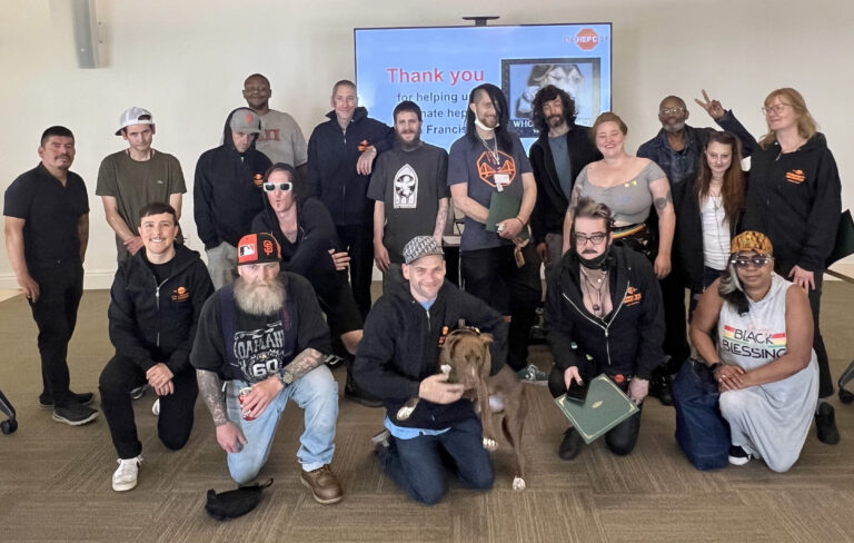 A smiling group of people and a dog stand and kneel in front of a backdrop with the words "Thank you".